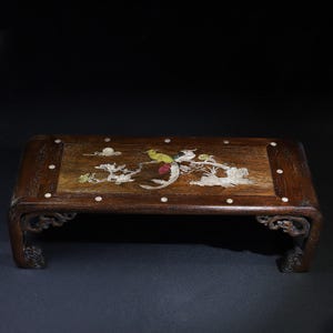 Può includere: Un tavolo rettangolare in legno con finitura marrone scuro. Il piano del tavolo presenta un disegno intarsiato di un uccello ed elementi floreali in vari colori. Il tavolo ha gambe curve con intagli decorativi e piccole intarsi a punti bianchi.