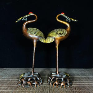 May include: A pair of antique bronze crane sculptures with gold accents. The cranes are standing on a base with intricate details. The cranes are facing each other with their heads tilted down.