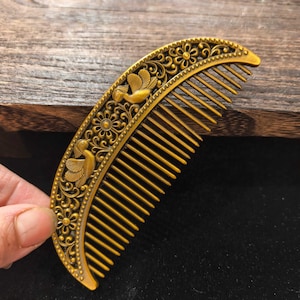 May include: Ornate gold-colored comb with a curved design. The comb features detailed floral and bird motifs along the top edge. The teeth of the comb are evenly spaced and the overall design is vintage-inspired.