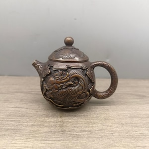 Puede incluir: Tetera de color bronce con un diseño detallado de dragón. La tetera tiene un asa curva y una tapa con un pequeño pomo. El dragón está intrincadamente tallado alrededor del cuerpo de la tetera.