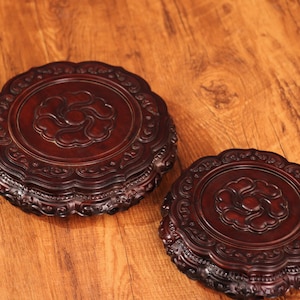 Puede incluir: Dos soportes redondos de madera de color rojo oscuro con diseños florales tallados y ornamentados. Los soportes tienen bordes festoneados y están colocados sobre una superficie de madera. Un soporte es más grande que el otro.