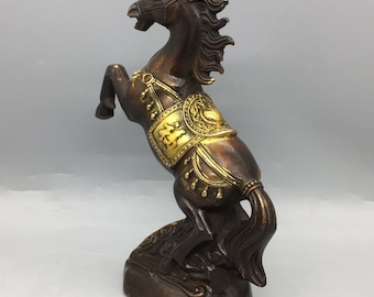 May include: A bronze-colored horse statue rearing up on its hind legs. The horse has a detailed mane and tail, with gold-colored accents on its saddle and harness. The statue is mounted on a rectangular base.