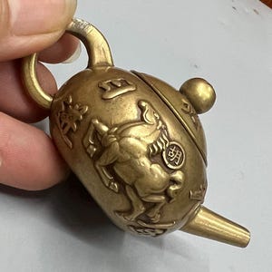 Puede incluir: Un pequeño colgante antiguo con forma de tetera de latón. El colgante presenta un relieve de un caballo y caracteres chinos. La tetera tiene un asa, un pico y una pequeña bola en la parte superior. El latón tiene una pátina cálida y envejecida.