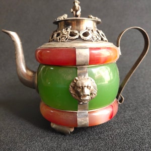 May include: A green and red jade teapot with silver accents and lion head details. The teapot has a silver lid with a small animal figure on top.