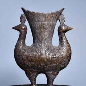 Könnte beinhalten: Eine bronzefarbene Metallvase in Form von zwei miteinander verbundenen Vögeln, die nach außen blicken. Die Oberfläche ist mit geprägten Federmustern, geschwungenen Ornamenten und strukturierten Schuppen auf Körpern, Hälsen und dem Rand verziert.