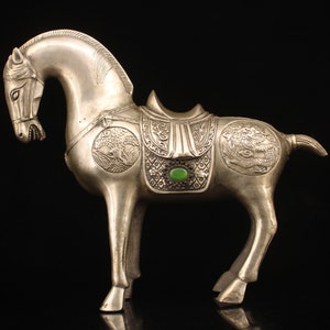 May include: A silver horse figurine with intricate details, including a saddle with a green gemstone. The horse is standing on a black background.