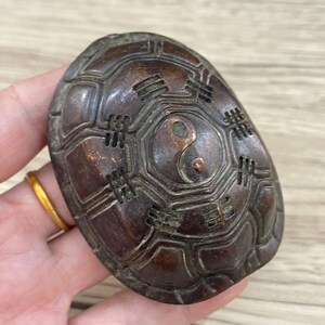 以下が含まれることがあります： 陰陽のシンボルと複雑な彫刻が施された、小さな濃い茶色の亀の甲羅。甲羅は金属製で、少し古びた外観をしています。