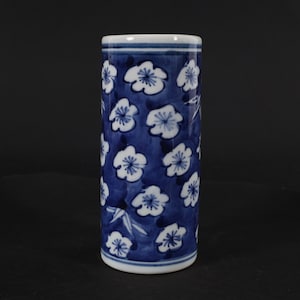 Op de afbeelding: Een witte keramische vaas met een blauw en wit bloemenpatroon. De vaas is cilindrisch van vorm en heeft een eenvoudig ontwerp.