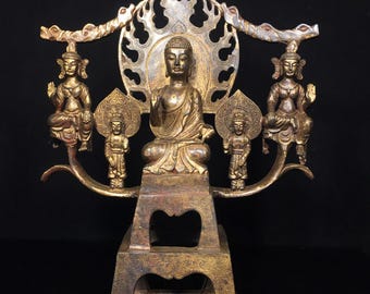 May include: A gold-colored metal sculpture of a seated Buddha with two figures on either side. The Buddha is surrounded by a decorative halo and sits on a raised platform.