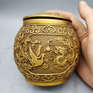 Könnte beinhalten: Ein goldfarbener Metallbehälter mit Deckel, der mit einem komplizierten Design aus Drachen und Phönix verziert ist. Der Behälter hat eine strukturierte Oberfläche und ist mit einem sich wiederholenden Wolkenmuster verziert.