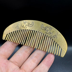 May include: A gold-colored metal comb with two elephant designs etched into the surface. The comb is curved and has a traditional design.