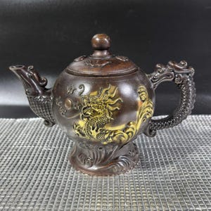 Op de afbeelding: Een bruine en gouden theepot met een drakenontwerp. De theepot heeft een deksel en een handvat in de vorm van een drakenstaart.