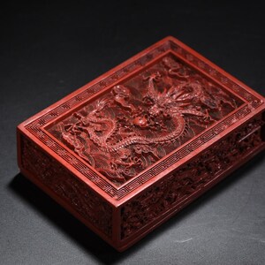 May include: A rectangular, red lacquered box with intricate dragon carvings on the lid and sides. The box features detailed relief work, showcasing a mythical dragon amidst swirling patterns. The box is a decorative item.