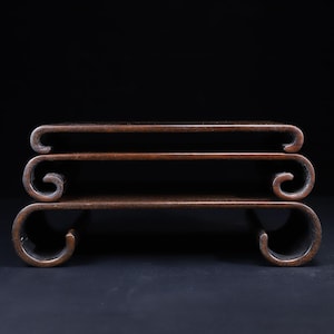 May include: A dark brown wooden display stand featuring a tiered design with three horizontal surfaces. Each level is supported by curved, scroll-shaped legs that extend outwards at the ends, creating a symmetrical and layered structure.