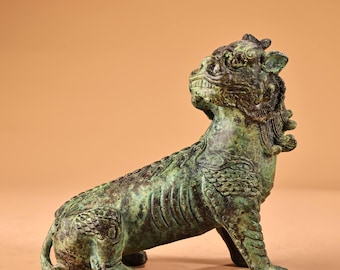 Puede incluir: Estatua de bronce de una criatura mítica con cuerpo de león y cabeza de dragón. La estatua presenta detalles intrincados, como escamas y pelaje, y una expresión feroz. La escultura está en posición sentada, mirando hacia arriba.