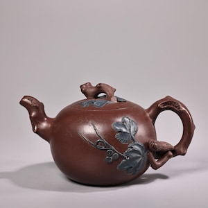 May include: A reddish-brown ceramic teapot with a rounded body, a curved spout, and a handle. The teapot is decorated with blue-grey leaf and vine designs. A small bird is perched on the handle. The lid has a small, decorative handle.