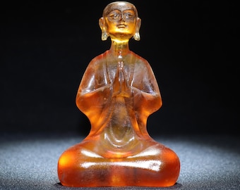 May include: A small, amber-colored glass figurine of a person in a seated meditation pose with hands clasped in front of their chest.
