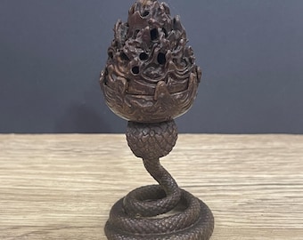 May include: A detailed, brown incense burner with a unique design. The burner features a flame-like top with intricate cutouts, a round midsection, and a snake-shaped base. The coiled snake base adds a distinctive touch to this decorative piece.