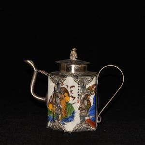 May include: A small, ornate teapot with a white body featuring a colorful painted scene. The teapot has a silver lid, handle, and spout. A small figurine sits atop the lid. The painted scene includes figures and text. The teapot is set against a black background.