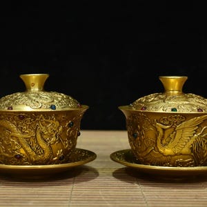 May include: Two ornate gold-colored tea cups with lids and saucers. Each cup features detailed relief carvings of dragons and birds, accented with colorful jewels. The cups are displayed on a light brown surface against a dark background.