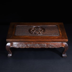 May include: A rectangular dark brown wooden display stand featuring a central circular relief carving surrounded by a textured grid pattern on the top surface. The apron and four curved legs are decorated with intricate carved scrollwork details.