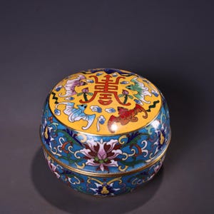 Op de afbeelding: Een ronde, decoratieve cloisonné doos met een gele deksel met Chinese karakters en kleurrijke vleermuisontwerpen. De doos heeft een blauwe basis met bloempatronen en gouden accenten. Het ontwerp is gedetailleerd.