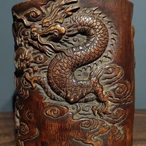 Puede incluir: Un cilindro de bambú marrón oscuro con un diseño de dragón tallado. El dragón está representado en una pose dinámica y en espiral, con la cabeza girada hacia un lado y la cola enroscada alrededor de su cuerpo. El fondo está lleno de nubes en espiral.