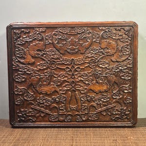 May include: A rectangular, wooden box with intricate carvings. The dark brown surface features detailed relief patterns of mythical creatures, clouds, and floral motifs. The box has a raised border and a rich, aged appearance, suggesting a vintage or antique origin.