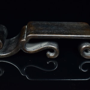 May include: A dark bronze decorative stand with a rectangular top and scrolled, curved supports. The stand features a rolled element on one end and a mirrored reflection below.