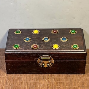 May include: A rectangular wooden box with a dark brown finish. The box is adorned with colorful gemstones set in gold-colored frames. A brass clasp secures the front. The box is likely used for jewelry or keepsakes.