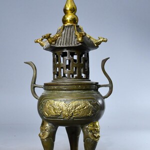 May include: A bronze incense burner featuring a pagoda-style lid with a gold-toned finial and dragon accents. The body displays relief carvings of dragons, two curved handles, and three legs adorned with sculpted lion-like faces in gold tones.
