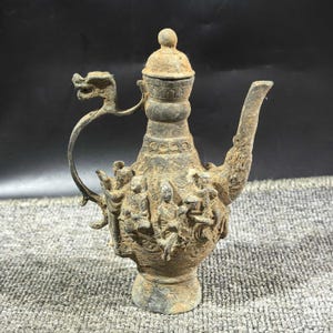 May include: Antique bronze-colored teapot with a dragon-shaped handle and spout. The body of the teapot is adorned with detailed relief carvings of figures. The lid is topped with a small knob. The teapot has a weathered appearance, suggesting age and use.