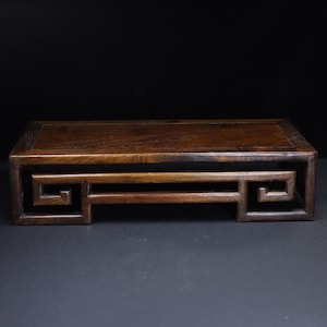 Può includere: Un espositore rettangolare in legno marrone scuro con un piano incassato e ritagli geometrici decorativi sui lati. L'espositore è lungo circa 30 cm e largo 15 cm, con uno sfondo scuro.