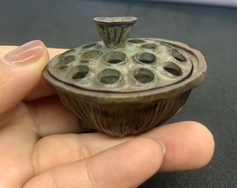 May include: A bronze-colored, lotus-shaped incense burner. The lid features multiple circular openings and a small handle. The burner is held in a hand against a dark background. The overall design is detailed and ornate.