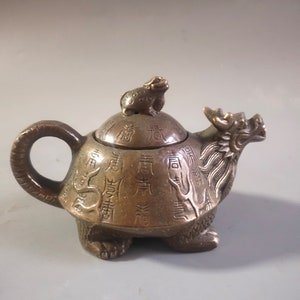 Puede incluir: Una tetera de bronce pequeña con forma de tortuga con una rana en la parte superior de la tapa. La tetera está decorada con caracteres chinos.