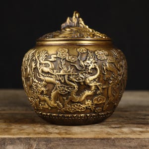 May include: A bronze-colored, round jar with a lid, featuring intricate relief carvings of dragons amidst clouds. The jar has a textured surface and sits on a wooden surface. The lid is topped with a mountain-like design.