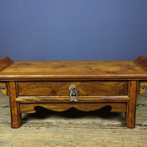 May include: A low rectangular wooden table with a rich brown grain, featuring a single central drawer with a decorative metal pull. The table has curved, flared ends on the top surface and a scalloped apron design between the four sturdy legs.