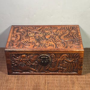 May include: A rectangular wooden chest with intricate dragon carvings. The chest is a rich brown color and features a metal clasp. The carvings depict dragons amidst swirling waves, adding a detailed, artistic element to the storage box.