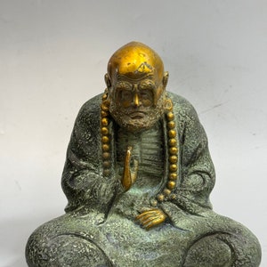 Puede incluir: Estatua de bronce de un hombre mayor calvo sentado en meditación. La estatua tiene la cabeza y las manos doradas, con un collar de cuentas. El hombre lleva una túnica y tiene las piernas cruzadas.
