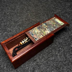 May include: A wooden box with a hinged lid, the lid is open and reveals a small compartment lined with iridescent mother of pearl. A black cord with a dark brown and white bead is inside the box.