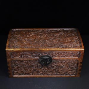May include: A rectangular wooden box with intricate dragon carvings on the lid and front. The box is a warm brown color with a dark metal clasp. The carvings depict dragons amidst clouds, showcasing detailed craftsmanship.