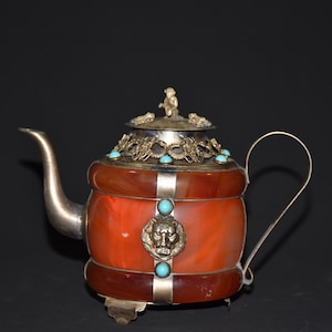 May include: A red carnelian teapot with silver and gold accents and turquoise stones. The teapot has a lion head design on the front and a small figure on the lid.