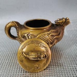 Puede incluir: Una tetera de color dorado con un diseño de dragón y una tapa con un diseño de pez. La tetera está hecha de metal y tiene un estilo chino tradicional.