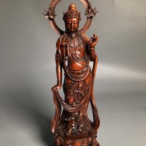 Op de afbeelding: Een donkerbruin houten beeld van een staande Boeddhafiguur met een halo en een vloeiende mantel. Het beeld is ingewikkeld gesneden en heeft een serene uitdrukking.