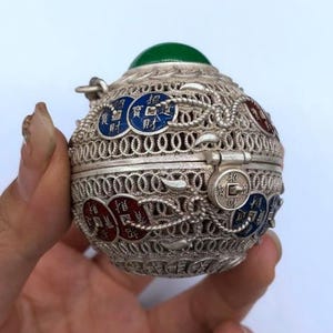 May include: A silver filigree orb-shaped container with red, blue, and white accents. The container has a hinged lid and a green gemstone on top. Decorative coins with Chinese characters are visible.