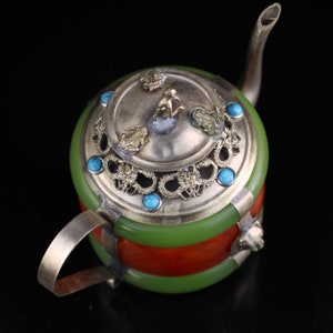 May include: A small, silver teapot with a green and orange jade body. The lid is decorated with a silver filigree design and turquoise stones. The teapot has a spout and a handle.