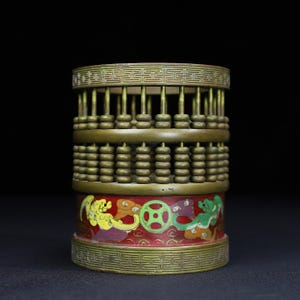 May include: A cylindrical brass abacus pen holder featuring a top and bottom rim with a geometric fretwork pattern. The upper section contains vertical rods with sliding beads, while the lower red base displays colorful painted dragon motifs.