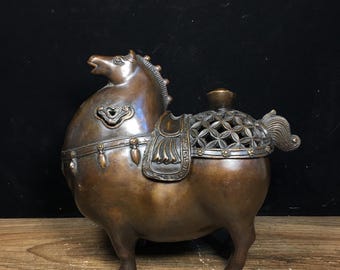 Puede incluir: Figura de caballo de bronce con detalles de silla de montar y un patrón floral en la espalda. El caballo está representado con la cabeza levantada, mostrando una melena y cola detalladas. La figura está sobre una superficie de madera sobre un fondo oscuro.