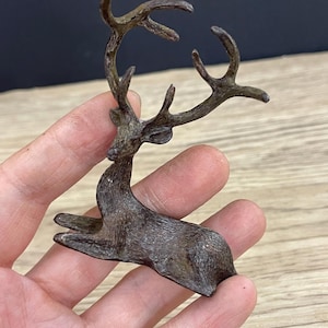 May include: A small, brown, metal deer figurine with antlers. The deer is sitting on its hind legs with its front legs outstretched.