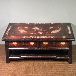 Può includere: Un mobile rettangolare in legno scuro con due cassetti e un ripiano inferiore. La superficie superiore presenta intarsi di pesci e motivi floreali. I cassetti hanno pomelli decorativi. Il mobile è di un ricco colore marrone-rossastro.